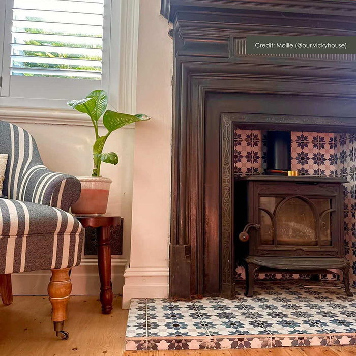 Living room interior featuring Mamounia River Blue 20x20cm Moroccan-style ceramic wall and floor tiles, paired with a cast iron stove, traditional fireplace, striped armchair, wall art, and soft neutral décor.