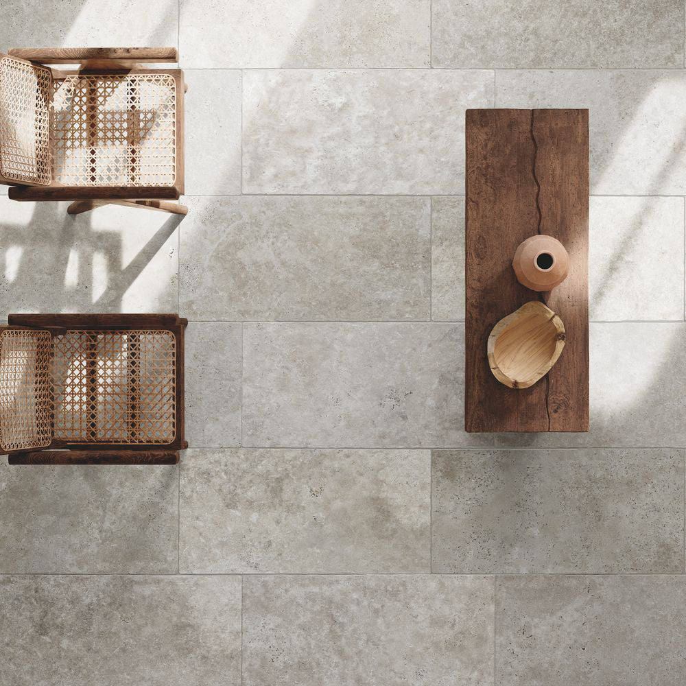 Monastry Flagstone XL Grey Tumbled Travertine 50 × 100cm porcelain floor tiles, styled with warm wood furniture and woven textures to highlight soft tonal variation, a gently aged finish, and a relaxed, natural stone effect.
