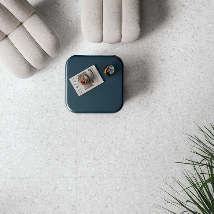 Top-down view of Noventa Blanco 3D Terrazzo 60x120cm porcelain floor tiles, showcasing a soft white terrazzo effect with subtle stone chips and textured 3D finish, paired with contemporary lounge furniture, a blue side table and indoor greenery.