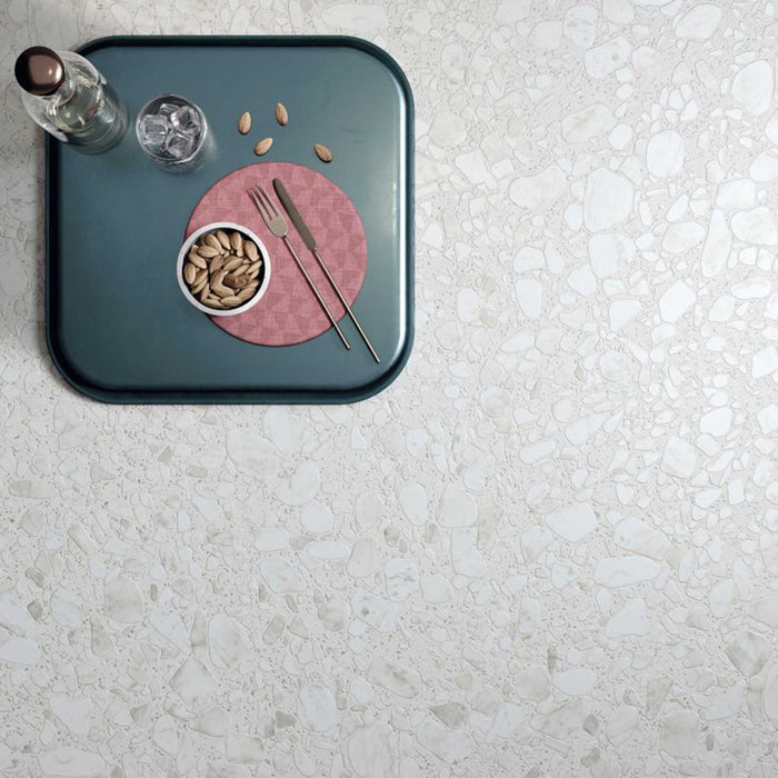 Top-down view of Noventa Blanco 3D Terrazzo 60x120cm porcelain floor tiles, showcasing a white terrazzo effect with large stone chips and textured 3D finish, styled with a blue serving tray, glassware, pistachios and modern tableware.