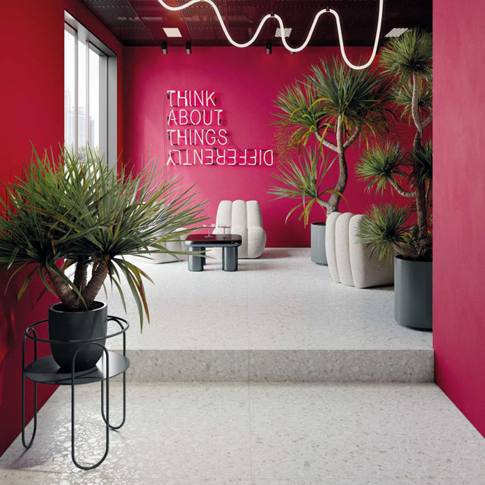 Commercial interior featuring Noventa Blanco 3D Terrazzo 60x120cm porcelain floor tiles, showcasing a white terrazzo effect with textured 3D stone chips, paired with bold pink walls, contemporary seating, statement lighting and tropical plants for a striking modern space.