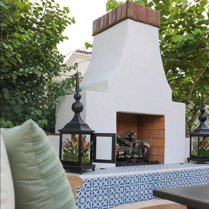 Outdoor fireplace setting featuring Iris Blue 20x20cm Victorian Moroccan-style patterned porcelain tiles, used as a decorative surround with crisp white masonry and greenery.