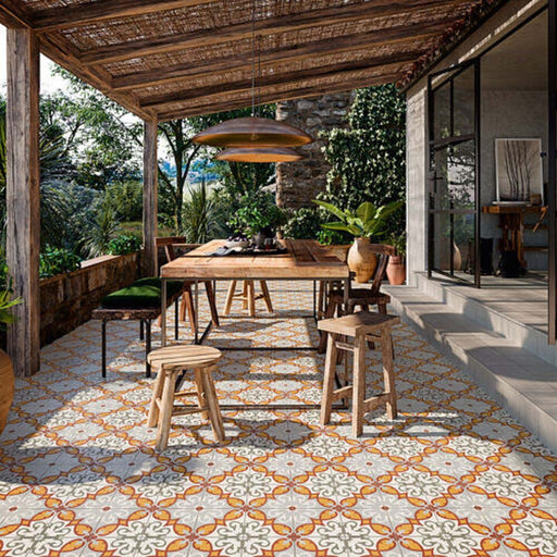 Rustic outdoor dining area featuring Palma Gela 2cm patterned outdoor slabs 50 × 100cm, showcasing a decorative Mediterranean-inspired pattern in warm terracotta, grey, and white tones, paired with natural wood furniture and lush greenery.