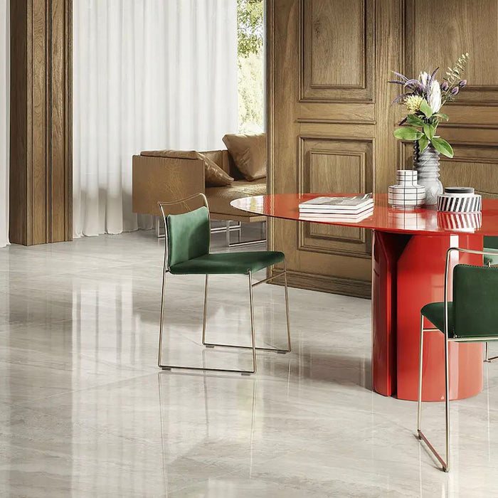 Living room featuring Paradis Marble Avorio Ivory stone marble effect porcelain wall and floor tiles in a 60cm x 120cm format, showcasing soft ivory, cream and light grey veining inspired by natural marble, paired with warm wood panelling, a bold red statement table, green upholstered dining chairs and contemporary furnishings for an elegant, design-led interior.