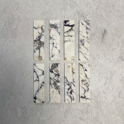 Eight Fitzrovia Picasso 5 x 20cm honed natural marble tiles arranged on a neutral backdrop, showcasing a light base with striking multi-tonal veining in deep charcoal, warm gold, and soft green tones, highlighting the natural variation of the stone.