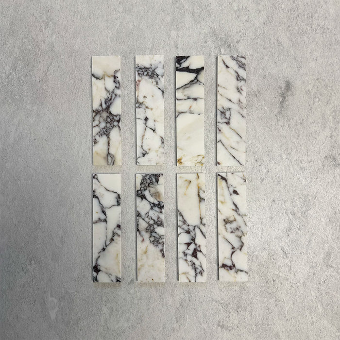 Eight Fitzrovia Picasso 5 x 20cm honed natural marble tiles arranged on a neutral backdrop, showcasing a light base with striking multi-tonal veining in deep charcoal, warm gold, and soft green tones, highlighting the natural variation of the stone.