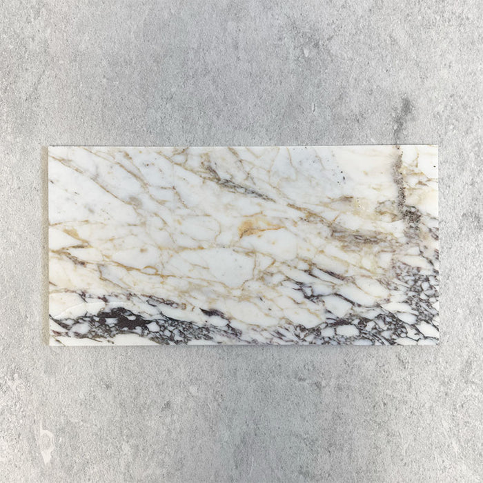Fitzrovia Picasso 30.5x61cm natural marble tiles with a honed finish, featuring a white base with intricate gold and grey veining, shown as a single tile sample laid horizontally against a light textured backdrop.
