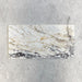 Fitzrovia Picasso 30.5x61cm natural marble tiles with a honed finish, featuring a white base with intricate gold and grey veining, shown as a single tile sample laid horizontally against a light textured backdrop.