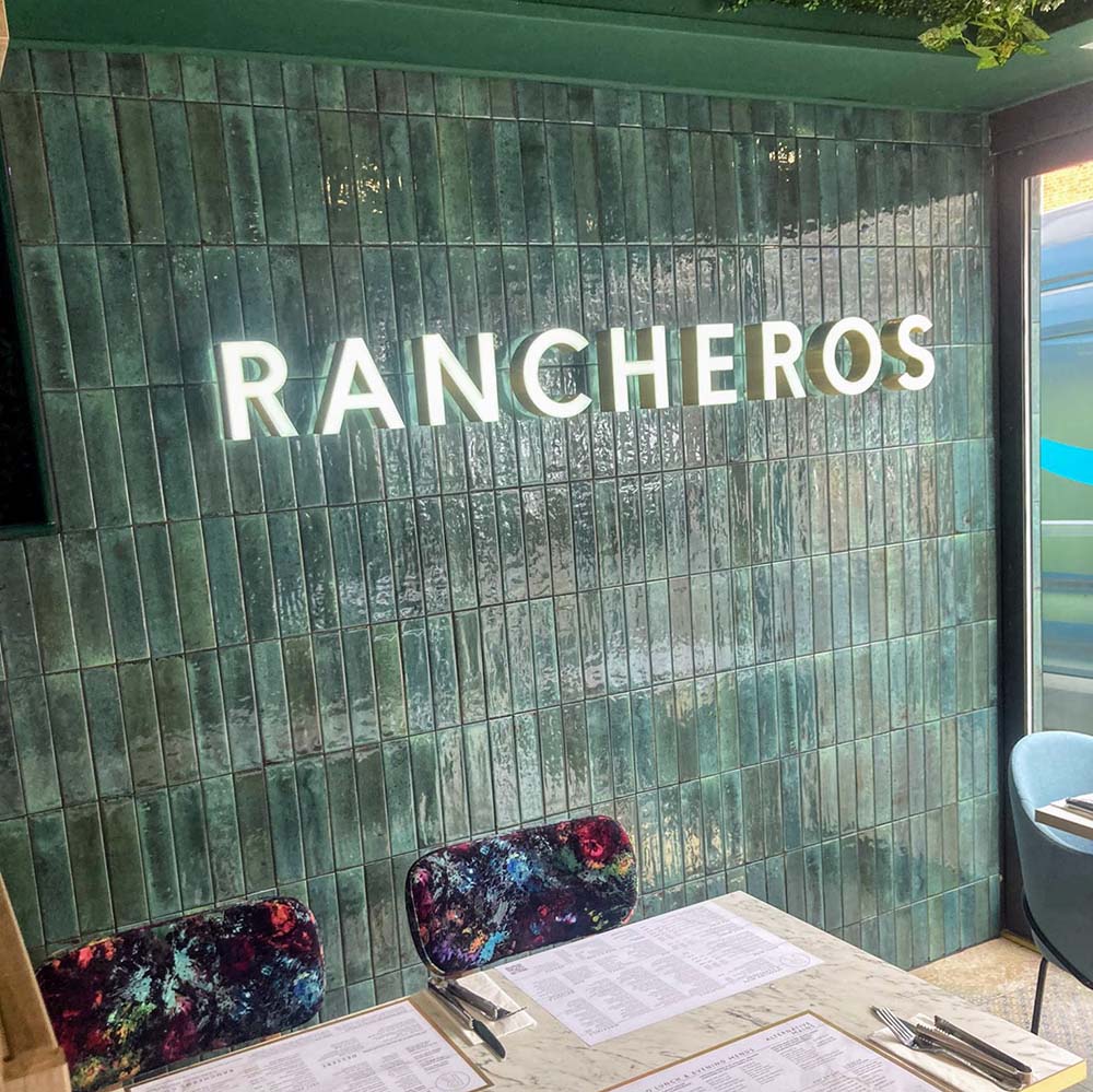 Restaurant interior featuring Izmir Brick Green Gloss 6x25cm ceramic wall tiles, styled with illuminated signage, marble tables, and vibrant patterned chairs for a bold, modern dining look.