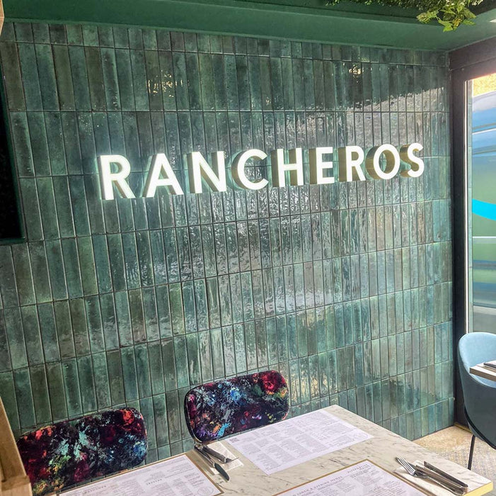Restaurant interior featuring Izmir Brick Green Gloss 6x25cm ceramic wall tiles, styled with illuminated signage, marble tables, and vibrant patterned chairs for a bold, modern dining look.