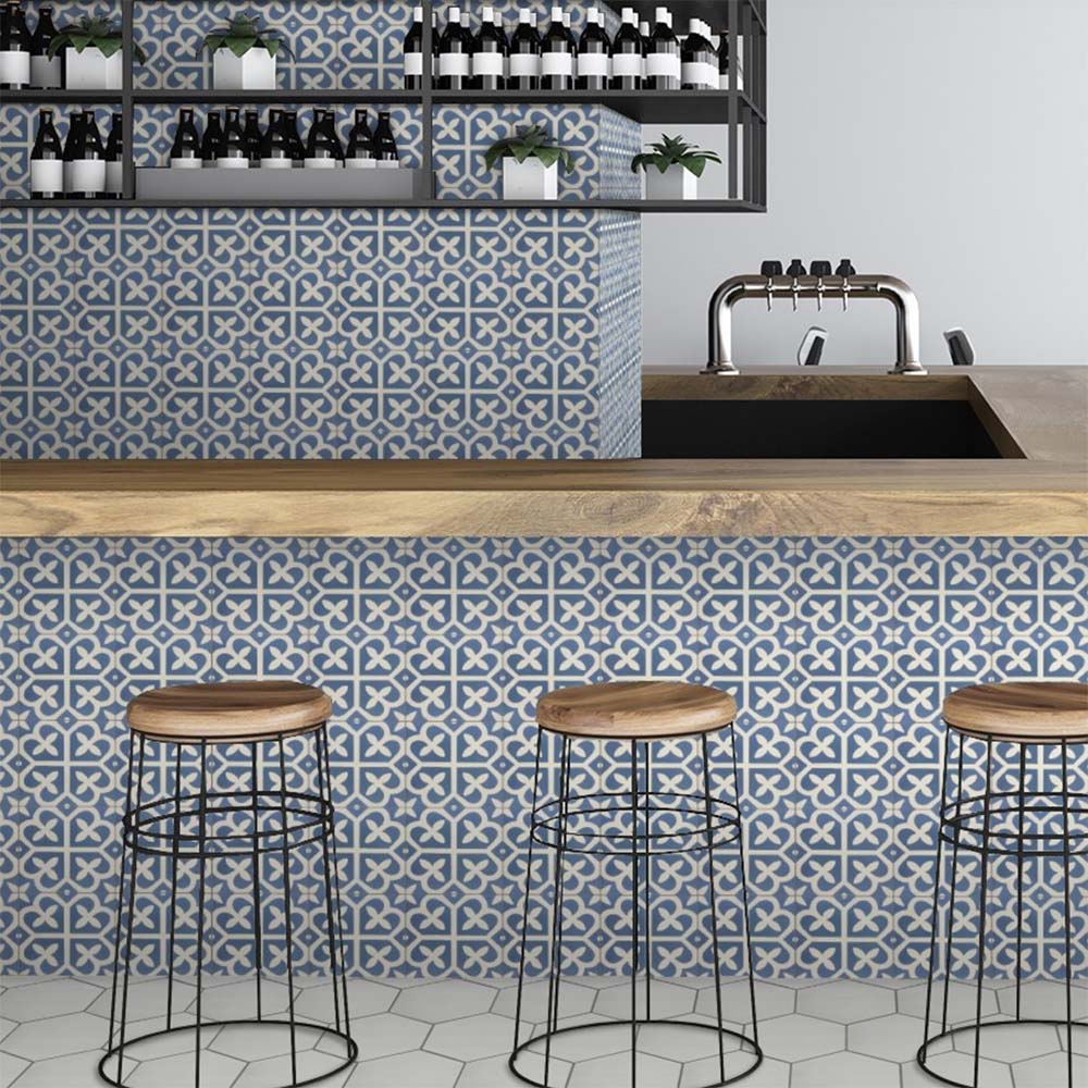 Restaurant interior featuring Iris Blue Victorian Moroccan-style 20×20cm porcelain tiles, used on bar fronts and walls to showcase a soft blue and cream geometric pattern, paired with wooden countertops, metal bar stools, and contemporary shelving.