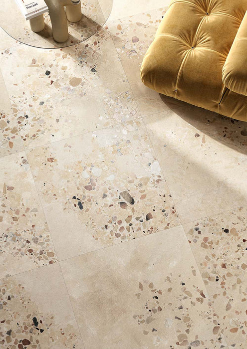 Living space featuring Riminy Sand Cosmo Stone wall and floor tiles 60 × 120cm, designed with a warm terrazzo-effect surface, scattered stone fragments, and soft sand-toned concrete detailing, paired with mustard velvet seating for a refined contemporary interior.