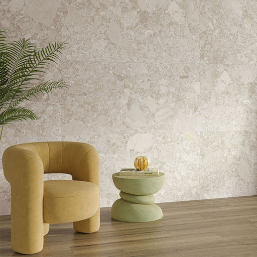 Living room interior featuring Contract Pebble Rock Avorio 60x120cm terrazzo effect porcelain wall tiles, styled with a curved mustard armchair, wooden flooring, a sculptural side table, and greenery for a warm, contemporary finish.