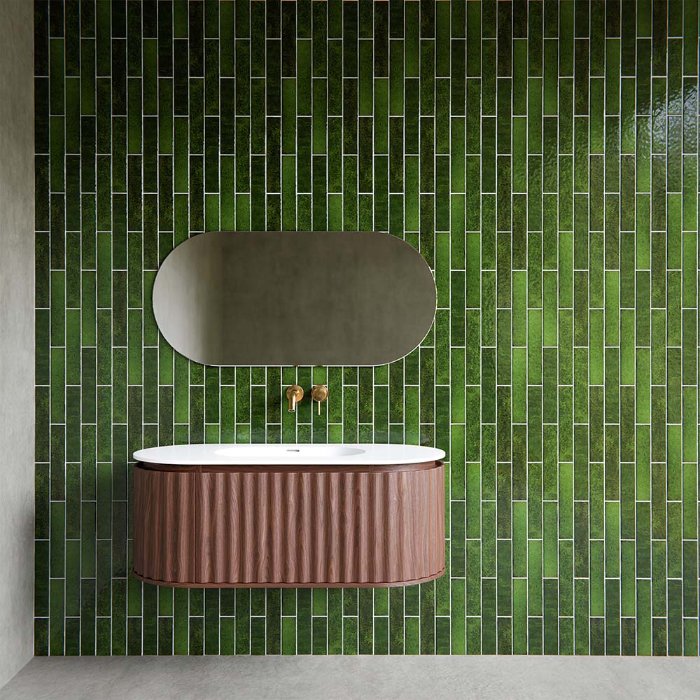 Malaga Tourmaline Green Brick 6.5cm x 32.8cm gloss wall tiles installed in a contemporary bathroom, featuring vertically stacked green brick tiles with tonal variation, paired with a curved wood vanity unit, oval mirror and brushed brass wall-mounted taps for a bold, design-led finish.