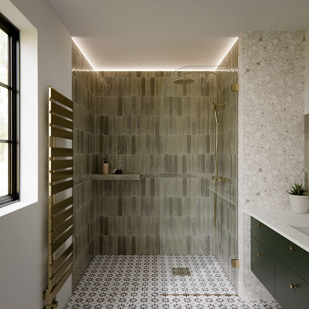 Izmir Sage Green 6x25cm gloss brick tiles used throughout a contemporary walk-in shower, showcasing soft sage tones with natural variation and a reflective glazed finish. The elongated format enhances vertical lines, adding texture and depth, while pairing beautifully with terrazzo-style walls, brass fittings and patterned floor tiles for a cohesive, design-led bathroom space.