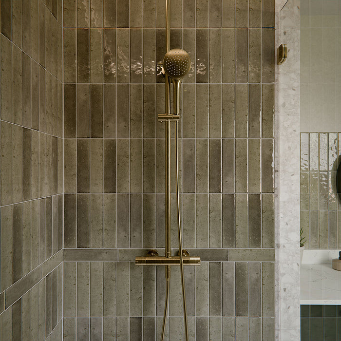 Izmir Sage Green 6x25cm gloss brick tiles used throughout a contemporary walk-in shower, showcasing soft sage tones with natural variation and a reflective glazed finish. The elongated format enhances vertical lines, adding texture and depth, while pairing beautifully with terrazzo-style walls, brass fittings and patterned floor tiles for a cohesive, design-led bathroom space.