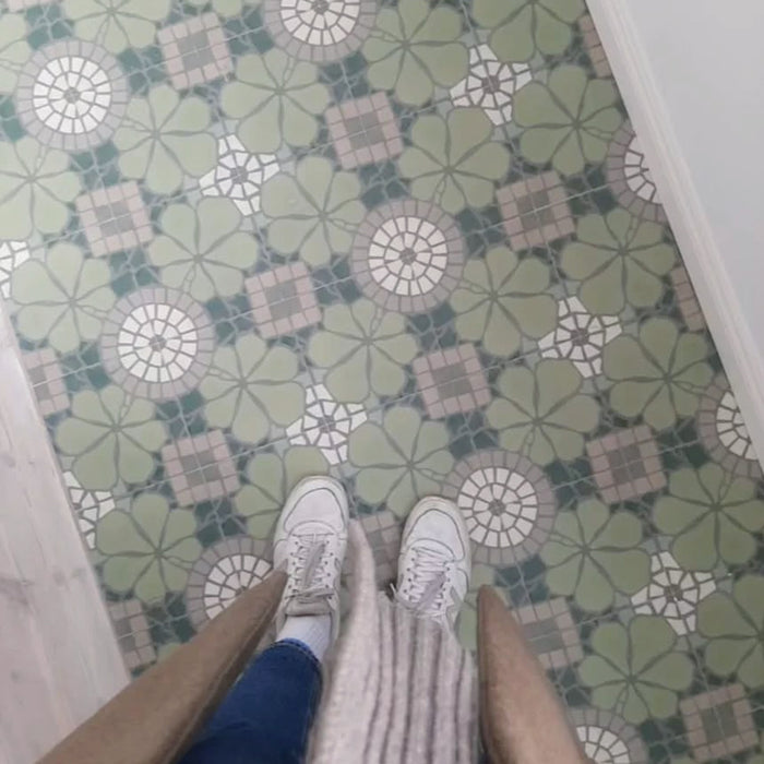 Overhead view of Bloom Green 20x20cm Moroccan-style ceramic floor tiles with green, white, and grey floral patterns, shown in a lifestyle setting with a person wearing white trainers and a coat standing on the tiles.