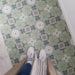 Overhead view of Bloom Green 20x20cm Moroccan-style ceramic floor tiles with green, white, and grey floral patterns, shown in a lifestyle setting with a person wearing white trainers and a coat standing on the tiles.