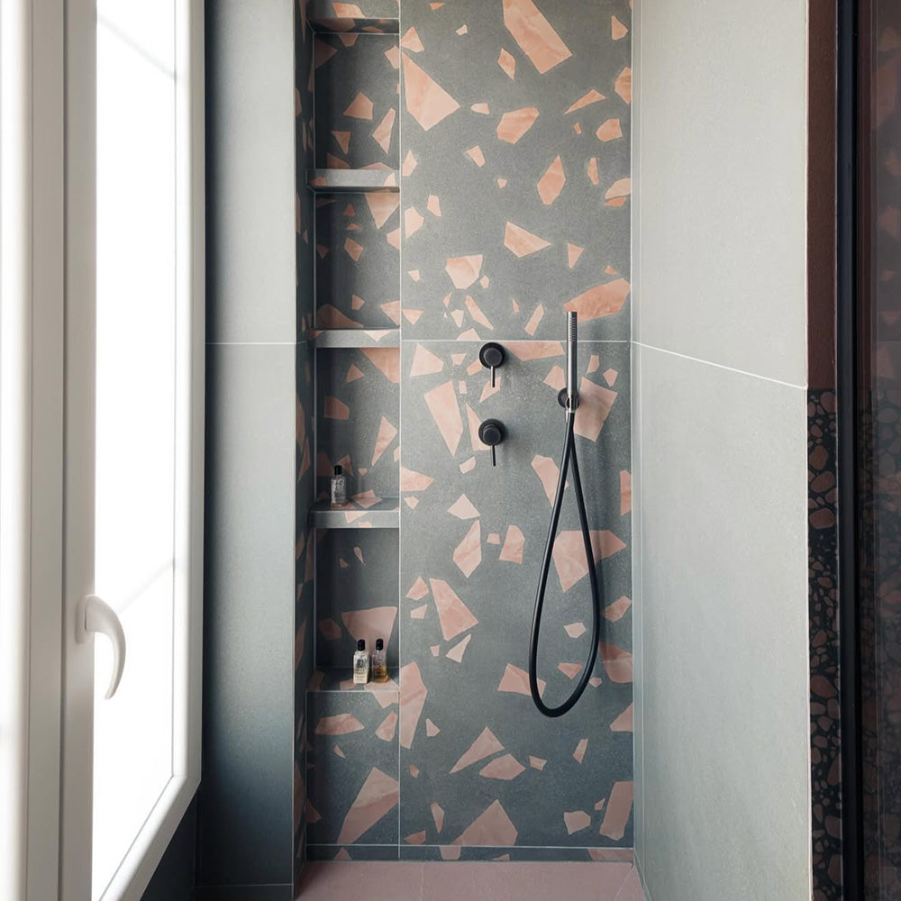 Shower enclosure featuring Chunks ROCK Terrazzo Green 60x120cm porcelain wall tiles, showcasing a terrazzo effect surface with warm pink and neutral fragments, paired with black fittings, built-in shelving, and a soft, contemporary bathroom setting.