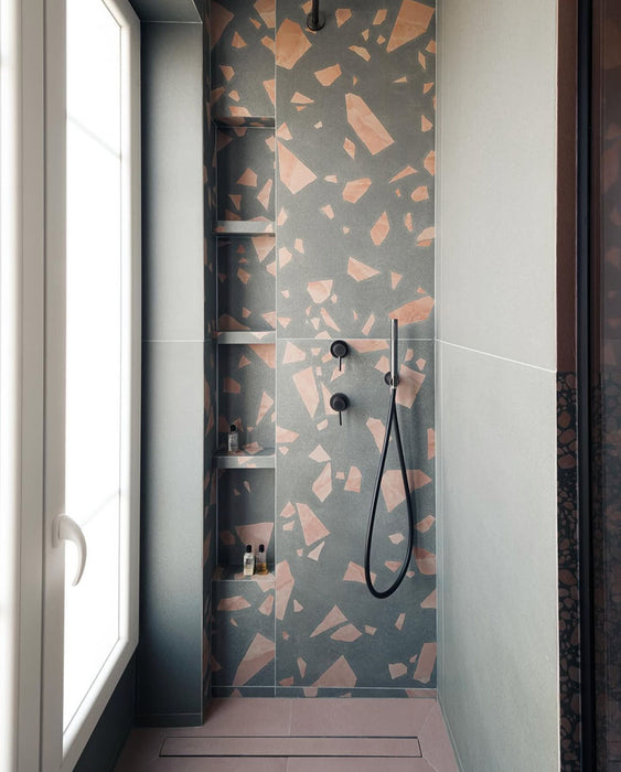 Shower enclosure featuring Chunks ROCK Terrazzo Green 60x120cm porcelain wall tiles, showcasing a terrazzo effect surface with warm pink and neutral fragments, paired with black fittings, built-in shelving, and a soft, contemporary bathroom setting.