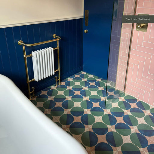 Bathroom featuring Geometry Ocean 20x20cm geometric patterned floor tiles in shades of blue, green, and sand, styled with a navy panelled wall, brass radiator, and pink herringbone shower tiles.