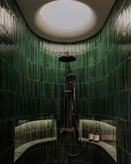 Curved shower area featuring Mizu green porcelain wall and floor tiles in a 6 × 24cm format, laid vertically to create a rich, glossy backdrop, complemented by built-in shelving and dark metal shower fittings.