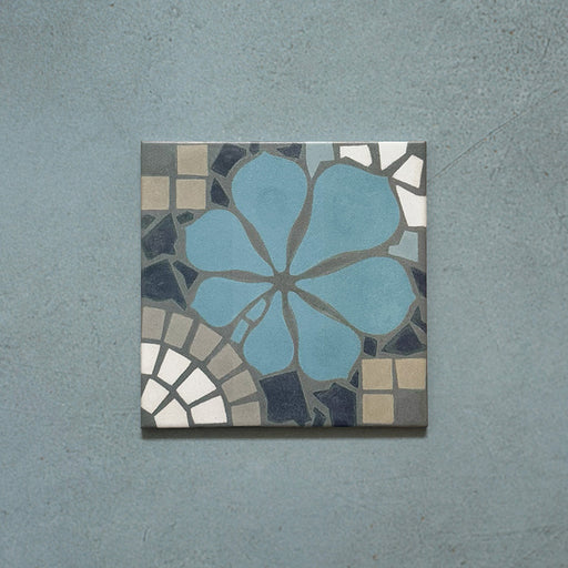 Single Bloom Azul Blue 20x20cm Moroccan-style ceramic tile featuring a floral pattern in blue, grey, beige, and white tones, shown against a textured blue background in a product close-up.