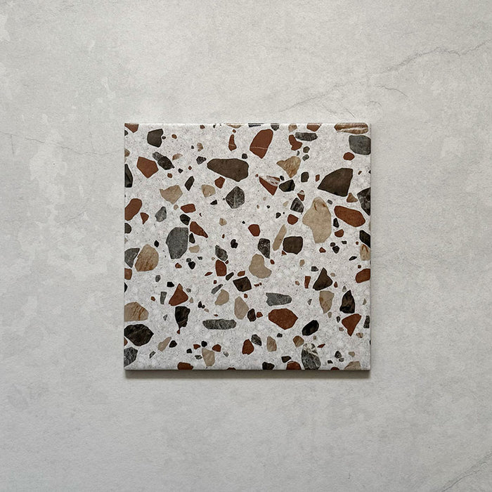 Flatlay featuring a single Dino Terrazzo Blanco Drops 20x20cm terrazzo-style ceramic tile, showcasing a white base with large stone-effect chips in earthy brown, terracotta, and grey tones.