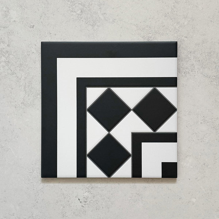 Single Sloane Square Black 25x25cm Victorian-style ceramic corner tile featuring a black and white geometric border and diamond pattern, shown against a light grey textured background in a product close-up.