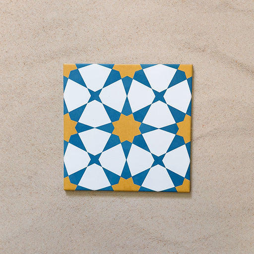 Close-up of Agraba Sunshine 20x20cm Moroccan-style ceramic tile featuring a bold geometric star pattern in blue, mustard yellow, and white on a neutral sandy background.