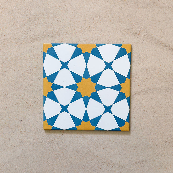 Close-up of Agraba Sunshine 20x20cm Moroccan-style ceramic tile featuring a bold geometric star pattern in blue, mustard yellow, and white on a neutral sandy background.