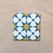 Close-up of Agraba Sunshine 20x20cm Moroccan-style ceramic tile featuring a bold geometric star pattern in blue, mustard yellow, and white on a neutral sandy background.
