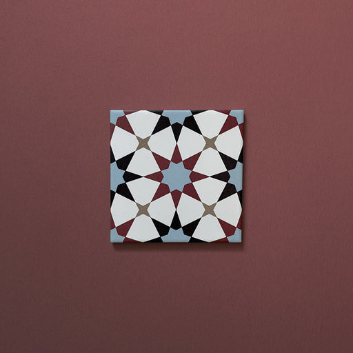 Close-up of Agraba Mini Atlas 15x15cm Moroccan-style ceramic tile featuring a geometric star pattern in blue, terracotta, black, and white tones on a warm burgundy background.