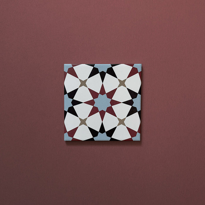 Close-up of Agraba Mini Atlas 15x15cm Moroccan-style ceramic tile featuring a geometric star pattern in blue, terracotta, black, and white tones on a warm burgundy background.