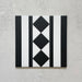 Single Sloane Square Black 25x25cm Victorian-style ceramic border tile featuring a black and white geometric stripe and diamond pattern, shown against a light grey textured background in a product close-up.