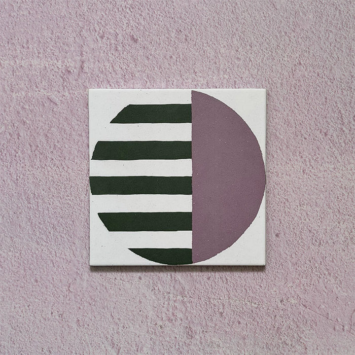 Single tile image showing the Geometry Seritina 20x20cm patterned tile with green horizontal stripes and a mauve semicircle on a cream base, photographed against a textured pink background.