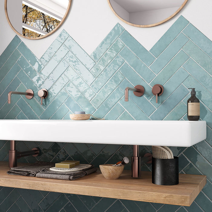 Madison Aquamarine Brick Wall Tiles 6x24.6cm — featured in a striking sink setting, these glossy teal tiles bring a fresh, ocean-inspired energy to the space. Their handcrafted finish and herringbone layout create texture and movement, beautifully complemented by warm copper fixtures and natural wood accents.