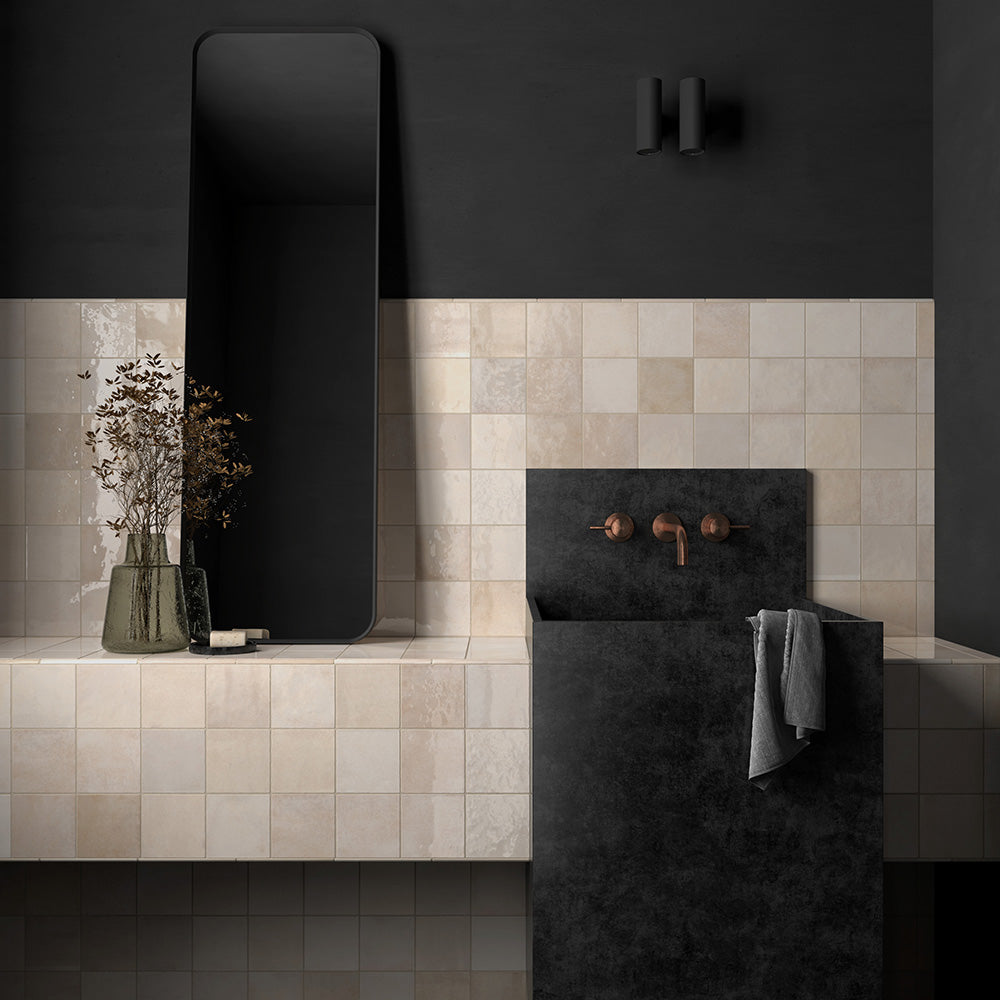 Bathroom sink setting featuring Zellij Bone Square Moroccan-style tiles in a 13.2cm x 13.2cm format, with warm beige tones and subtle handmade surface variation, styled with a dark stone basin, wall-mounted brass tapware, and a sleek black mirror.