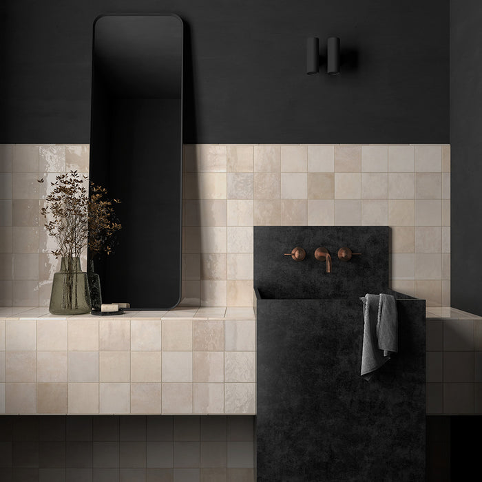 Bathroom sink setting featuring Zellij Bone Square Moroccan-style tiles in a 13.2cm x 13.2cm format, with warm beige tones and subtle handmade surface variation, styled with a dark stone basin, wall-mounted brass tapware, and a sleek black mirror.