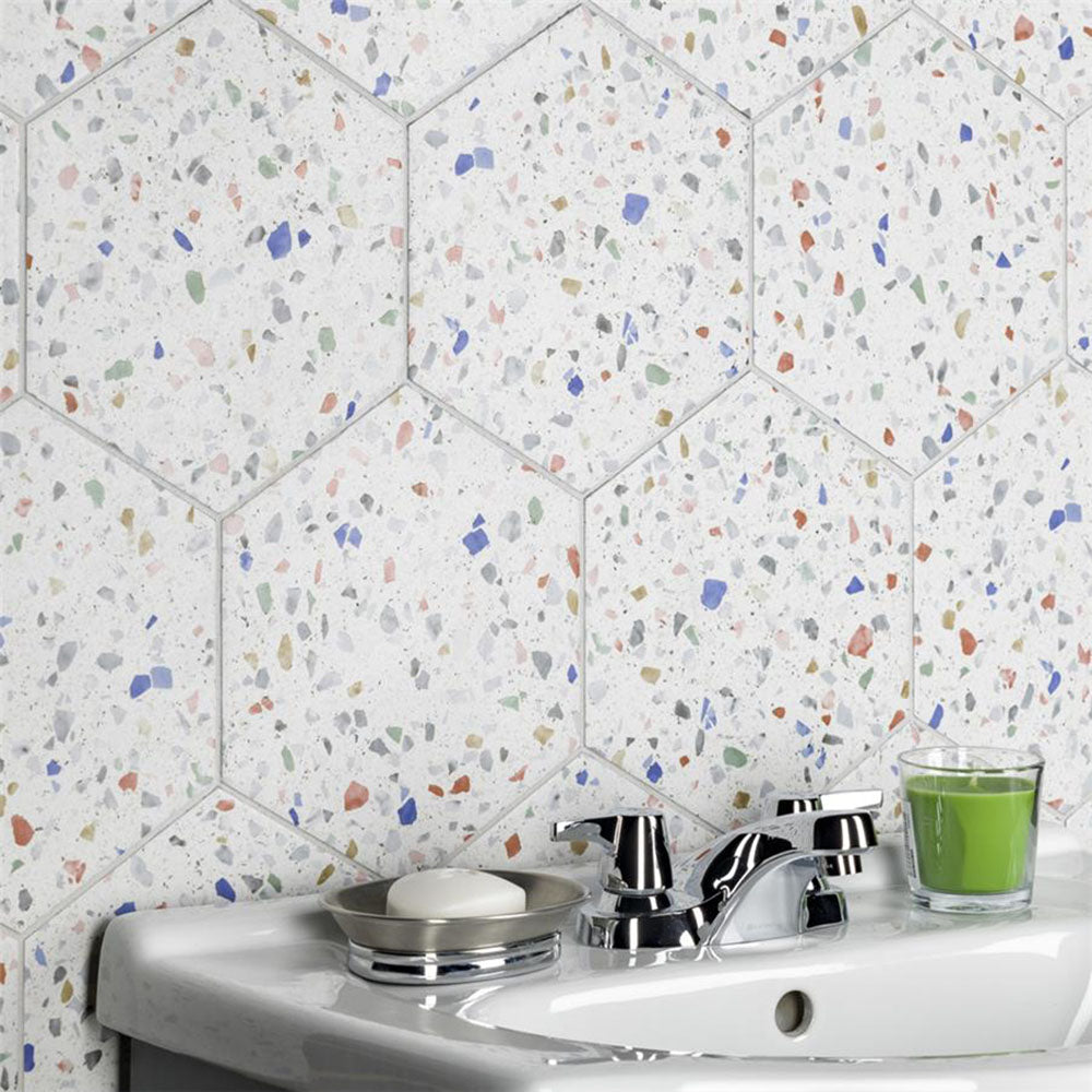 Bathroom sink setting featuring Confetti Tutti Frutti Hexagon 22×25cm terrazzo-style porcelain wall tiles with colourful multitone chips on a white base, creating a playful geometric backdrop behind a white basin with chrome fixtures.