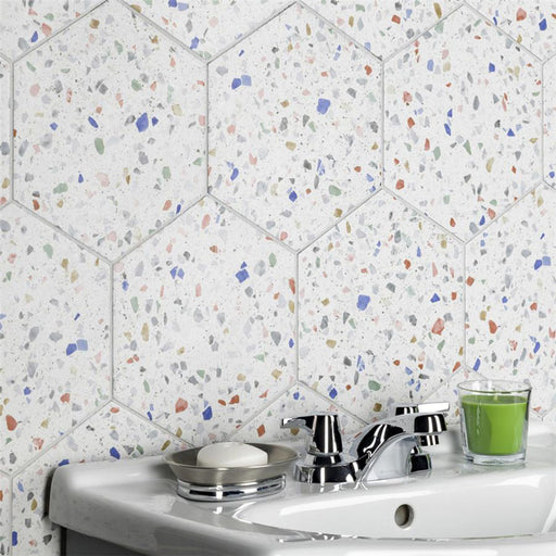 Bathroom sink setting featuring Confetti Tutti Frutti Hexagon 22×25cm terrazzo-style porcelain wall tiles with colourful multitone chips on a white base, creating a playful geometric backdrop behind a white basin with chrome fixtures.