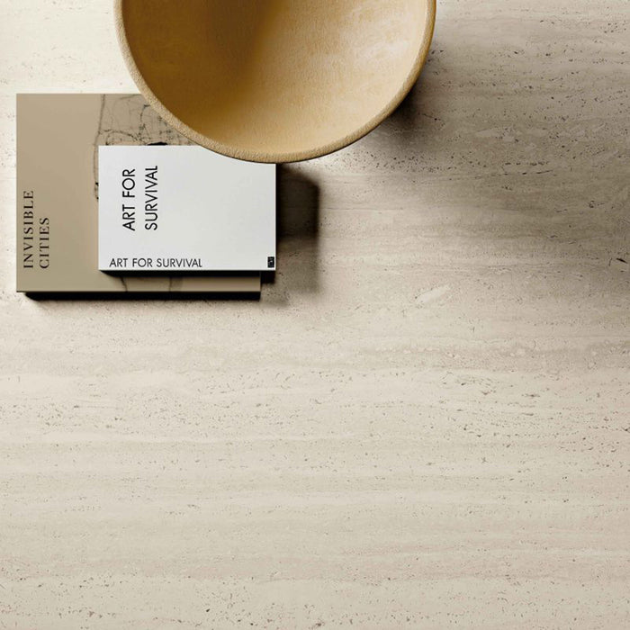 Close-up of Storico Travertine Stone matt porcelain tiles showcasing a soft travertine effect surface with delicate linear veining, styled with warm neutral décor and books to highlight the tile's natural, contemporary appearance.