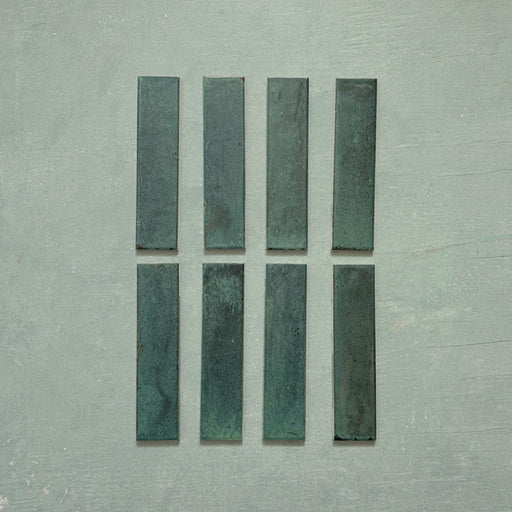 Eight Izmir Brick Green Gloss 6x25cm ceramic tiles displayed on a soft green background, showing rich tonal variation and a reflective glossy finish.