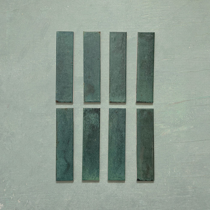 Eight Izmir Brick Green Gloss 6x25cm ceramic tiles displayed on a soft green background, showing rich tonal variation and a reflective glossy finish.