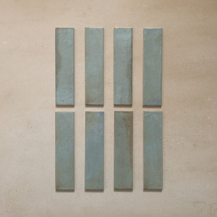 Eight Izmir Brick Celadon Green Gloss 6x25cm ceramic tiles displayed on a neutral beige background, showing subtle tonal variations and a smooth glazed finish.