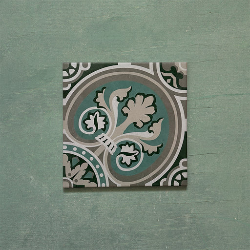 Single Europa Toulouse 20x20cm Victorian Moroccan style tile displayed against a textured green background, featuring an ornate floral and geometric pattern in green, grey and cream tones in a close-up product setting.