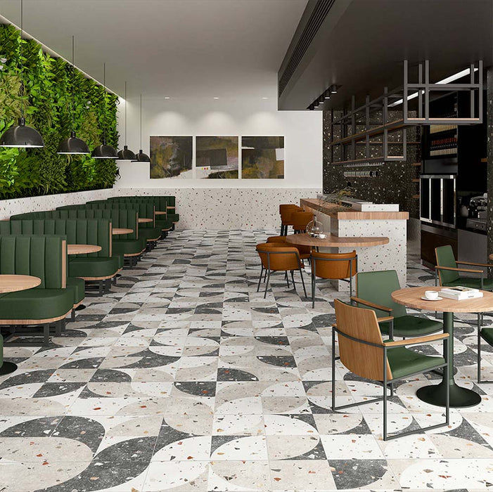 Contemporary café interior featuring Tulsa Decor 66 × 66cm terrazzo-effect porcelain wall and floor tiles, showcasing a bold geometric pattern in contrasting ivory and charcoal tones with scattered terrazzo fragments, paired with warm wood finishes, green seating, and statement lighting for a striking commercial space.