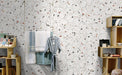 Retail-style interior wall featuring Tulsa White Terrazzo Hexagon 25 × 22cm porcelain tiles, with a crisp white base and scattered multicoloured aggregate chips, laid in a honeycomb pattern and paired with light wood shelving and soft textiles for a fresh, contemporary display.