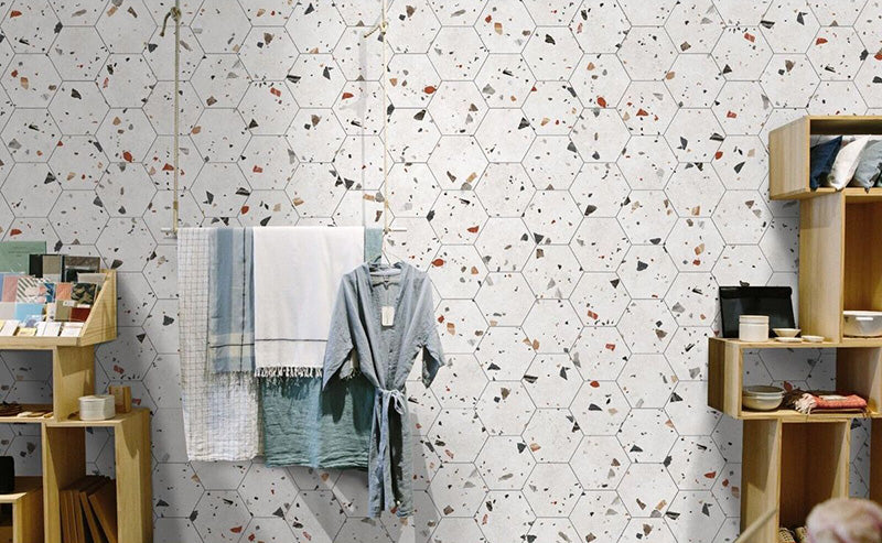 Retail-style interior wall featuring Tulsa White Terrazzo Hexagon 25 × 22cm porcelain tiles, with a crisp white base and scattered multicoloured aggregate chips, laid in a honeycomb pattern and paired with light wood shelving and soft textiles for a fresh, contemporary display.