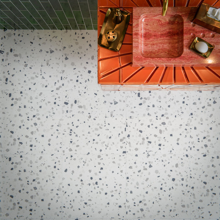 Bathroom interior featuring Veneto Medium White 60 × 60cm terrazzo-effect porcelain floor tiles, showcasing a soft white base with scattered grey stone fragments, paired with Malaga Tourmaline Green gloss brick wall tiles, a statement coral vanity unit, and brushed brass fittings for a bold yet balanced contemporary space.